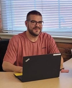 A man in a red shirt and black glasses sits at a laptop in the Albany office of New York Registered Agent LLC.